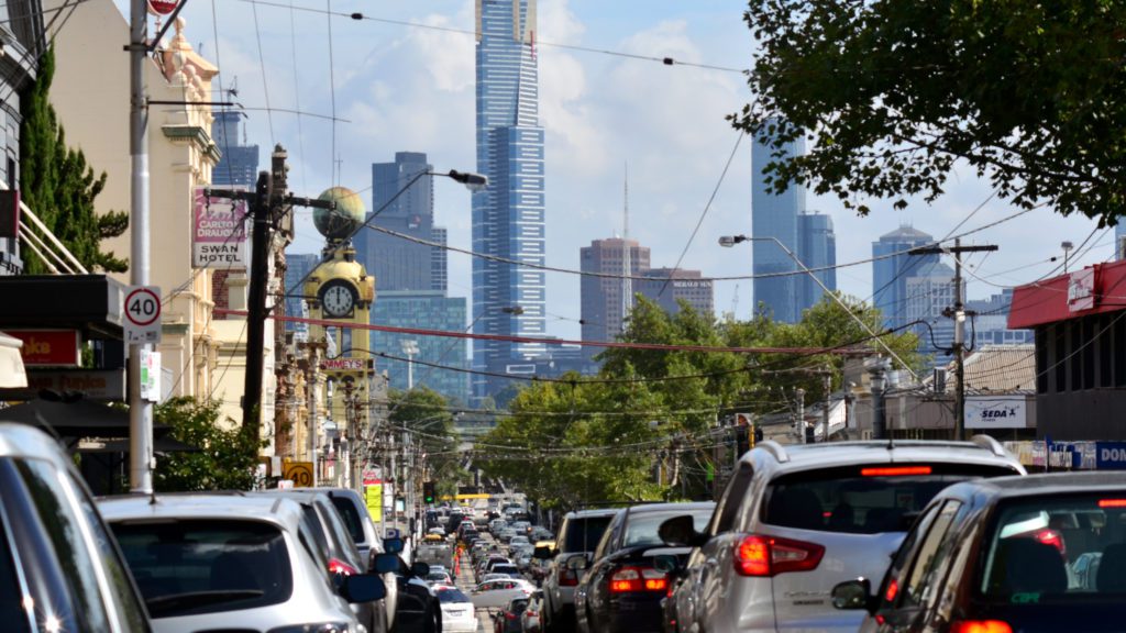 A long line of cars on a narrow, crowded street in Melbourne with skyscrapers ahead in the distance.